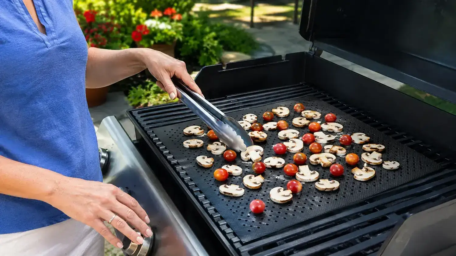 Hands placing cut vegetables onto a black non-stick grill mat resting on a charcoal grill grate